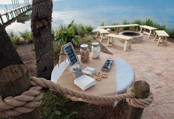 An outdoor dining area with a table set for a meal, surrounded by a rope barrier, with a view of the water and a wooden deck in the background.