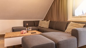 Ein modernes Wohnzimmer mit einem grauen Ecksofa, einem Couchtisch aus Holz mit Flasche und Glas sowie Blick auf eine Brücke durch ein Fenster.