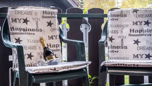 Zwei Gartenstühle mit Picknickausstattung, darunter eine Flasche und Gläser, vor einem Holzzaun mit einem Haus im Hintergrund.