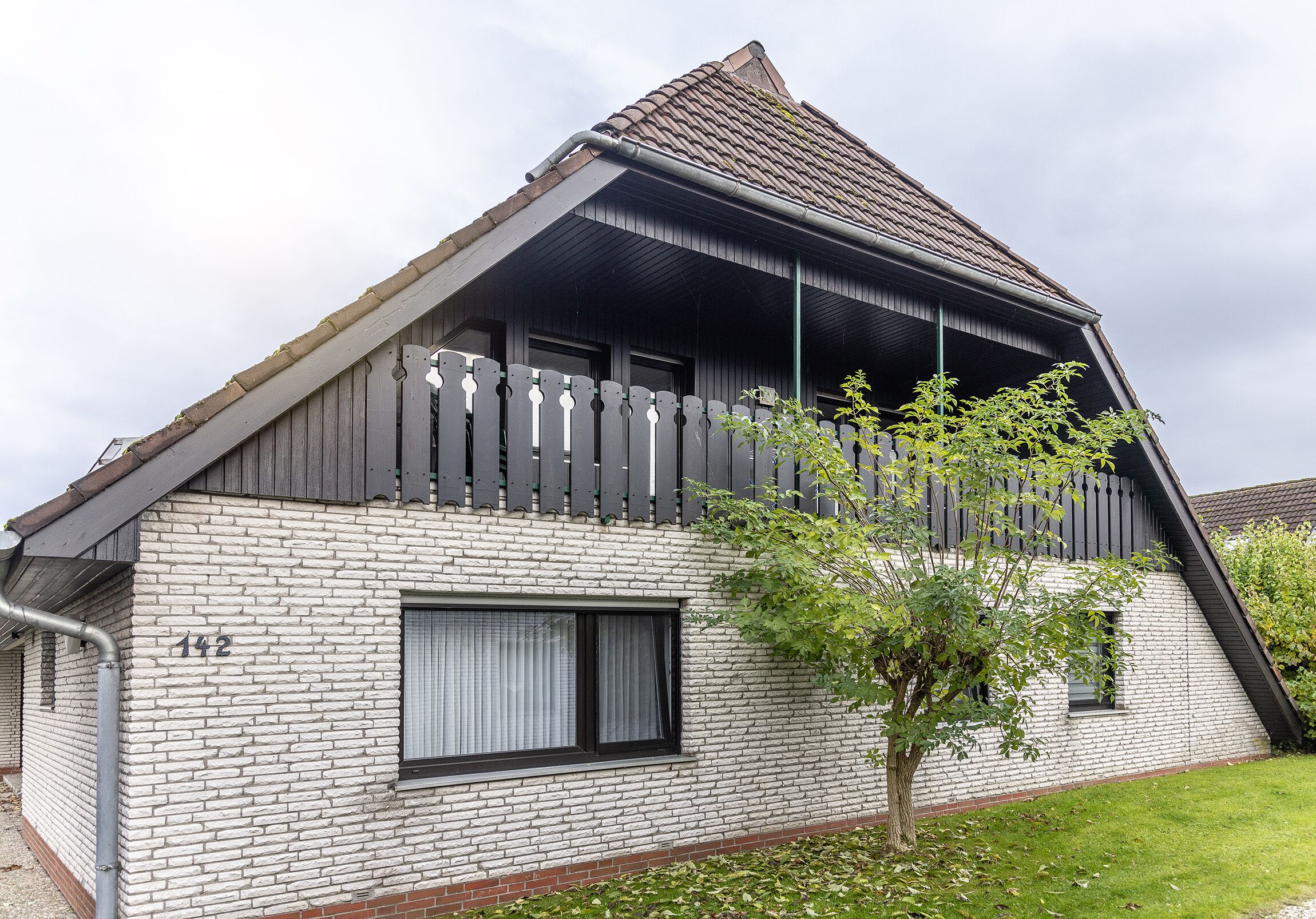 A house with a sloped roof, a balcony with a wooden railing, and a small window with curtains.
