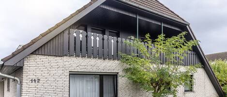 A house with a sloped roof, a balcony with a wooden railing, and a small window with curtains.