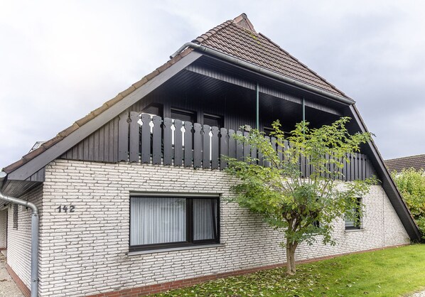 Une maison avec un toit en pente, un balcon doté d’une balustrade en bois et une petite fenêtre avec des rideaux.