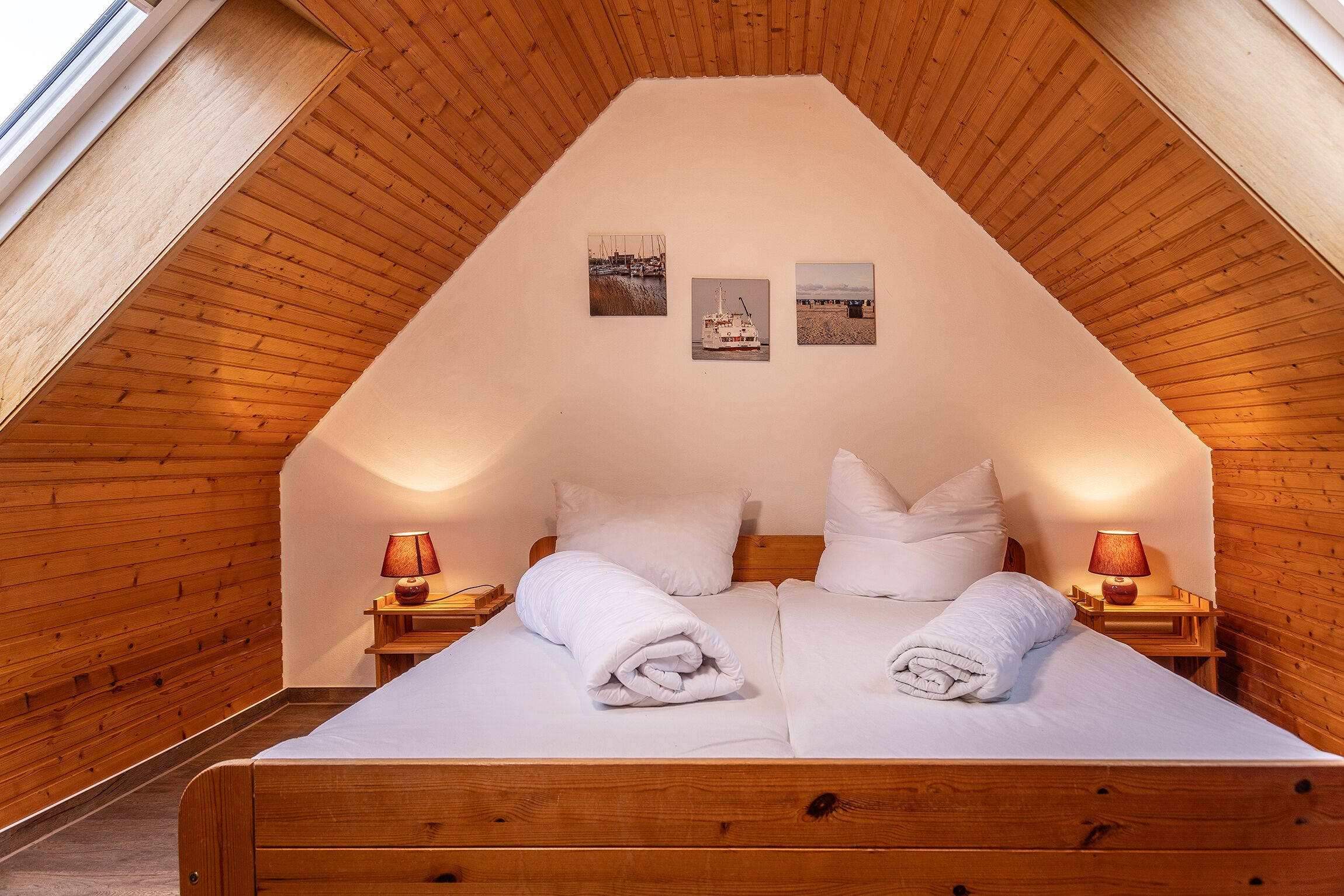 Een slaapkamer met een schuine kap, houten wanden, een bed met witte lakens, twee bedlampjes en drie ingelijste foto's aan de muur.