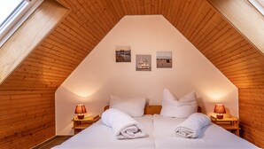 Ein Schlafzimmer mit geneigter Decke, Holzwänden, einem Bett mit weißen Leinen, zwei Nachttischlampen und drei gerahmten Bildern an der Wand.