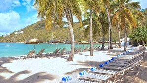 Ein Strand mit Reihen von Liegestühlen und Sonnenschirmen, ein klares blaues Meer und ein Hügel mit Gebäuden im Hintergrund.