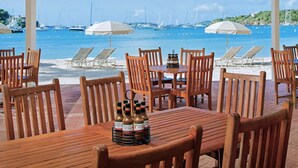 Ein Restaurant am Strand mit Holztischen und -stühlen, ein Tisch mit Flaschen und Sonnenschirmen mit Blick aufs Meer.