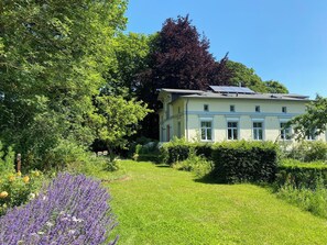 Ein zweigeschossiges Gebäude mit Garten, umgeben von Bäumen und verschiedenen Pflanzen.