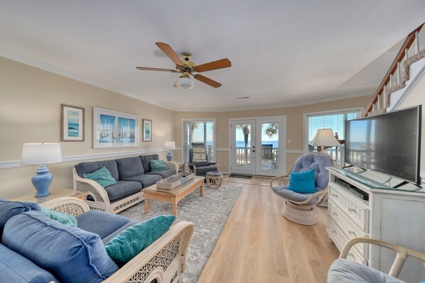 A spacious living room with a ceiling fan, a television, wicker furniture, and a view of the outdoors through large windows.