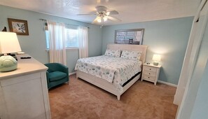 Ein Schlafzimmer mit einem Bett, einem Sessel, einem Nachttisch, einer Lampe, einem Deckenventilator und einem Fenster mit Vorhängen.