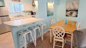 Een compacte keuken met een centraal eiland, witte barstoelen, een eettafel met stoelen en een schilderij van een zeilboot.