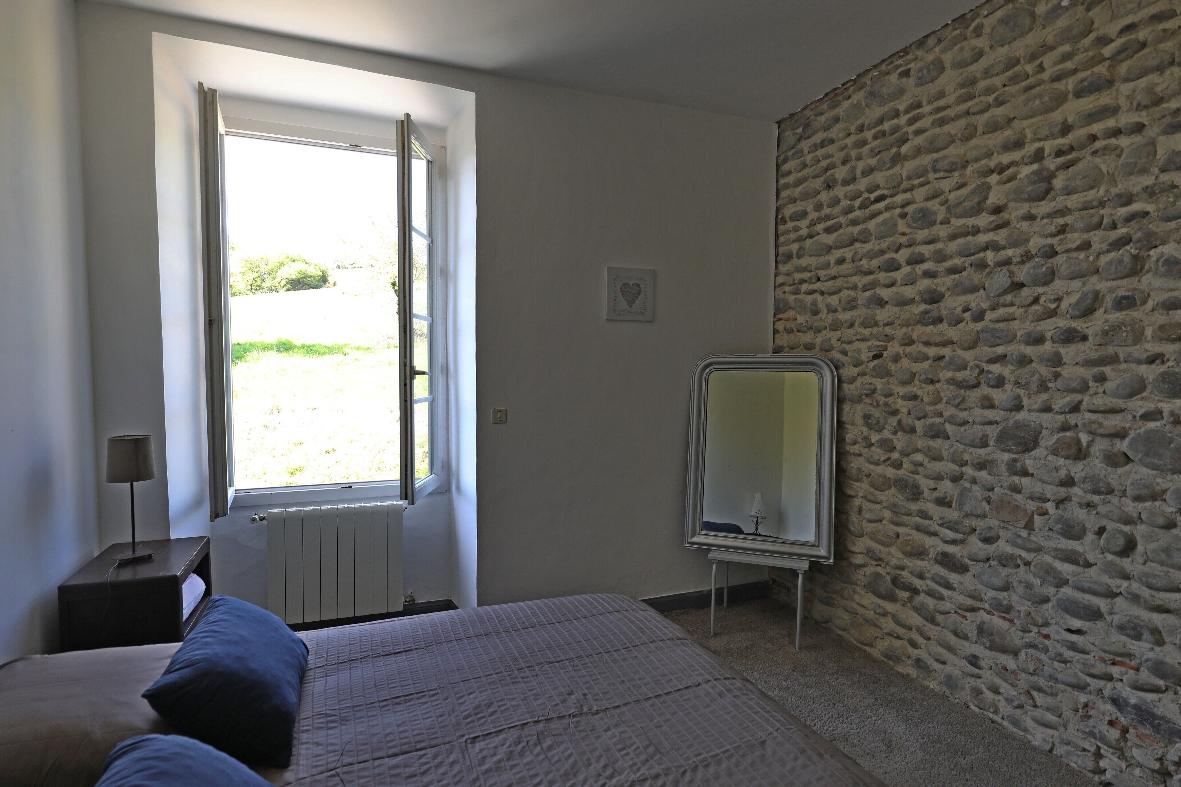 Une chambre à coucher comprenant un lit, une table de chevet, une commode, une fenêtre donnant sur un paysage agréable et un mur en pierre.