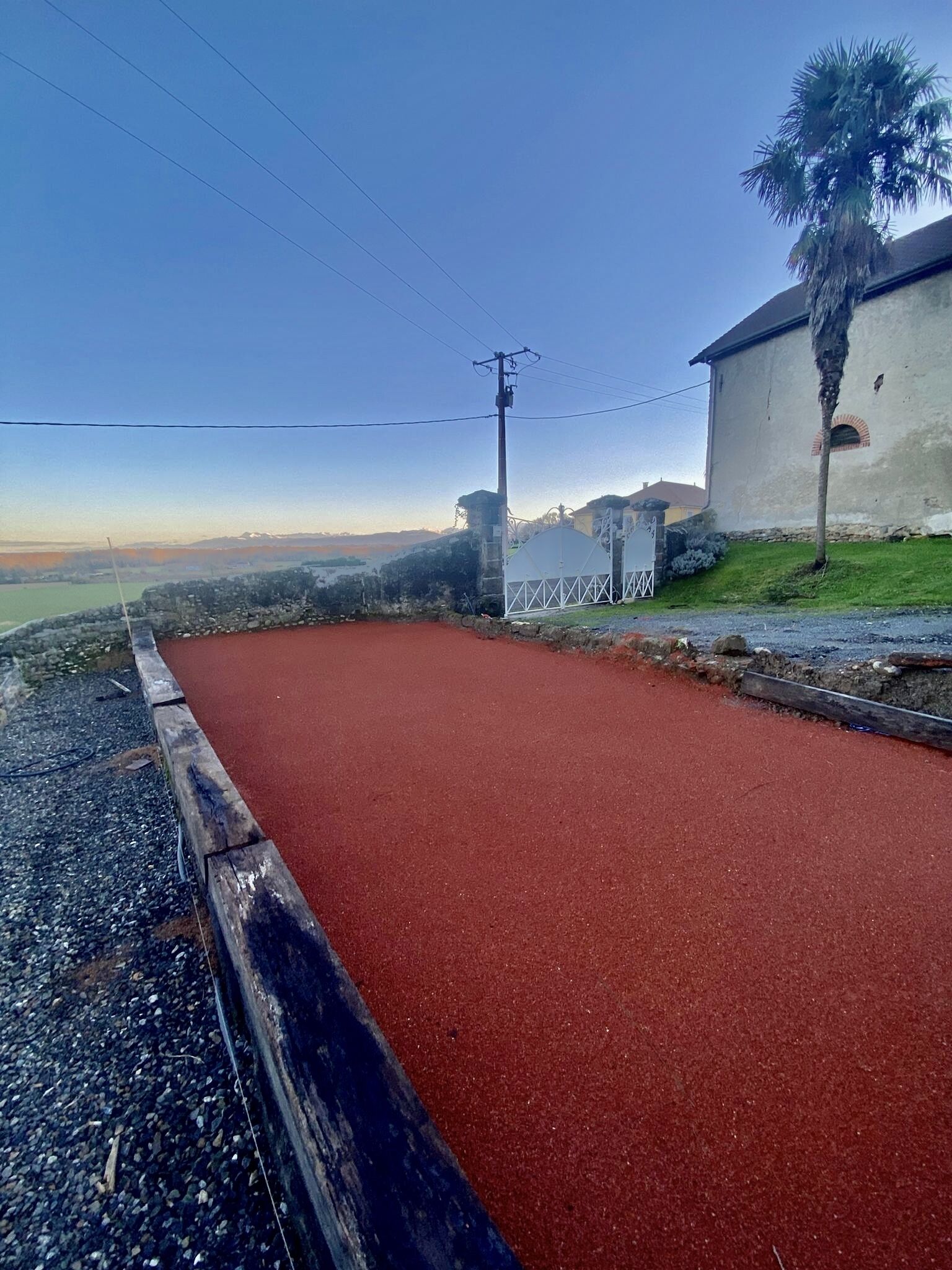 Un chemin pavé à la surface rouge, bordé d’un mur de pierre et d’un bâtiment à la façade blanche, avec un palmier.