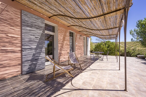 A covered outdoor area with a pergola, a table with chairs, and a view of a vineyard.