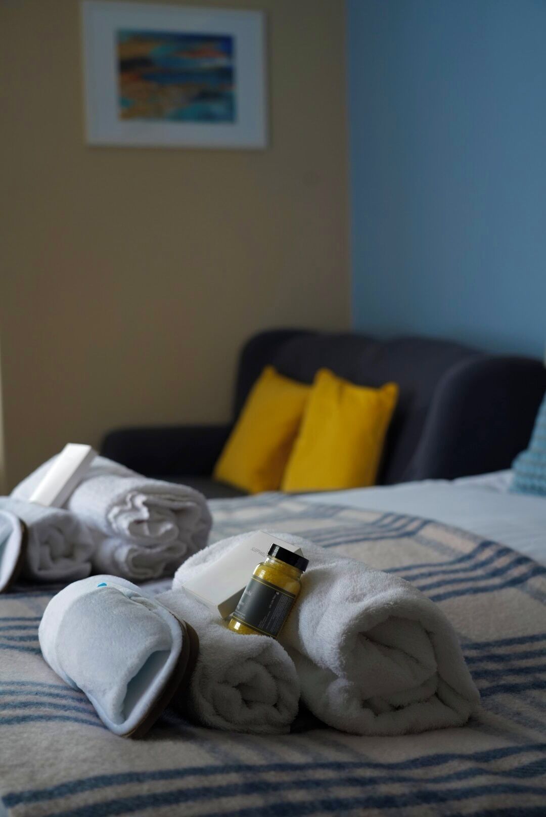 Una cama bien hecha con toallas enrolladas y una botella de loción, una silla azul oscuro con almohadones amarillos y un cuadro en la pared.