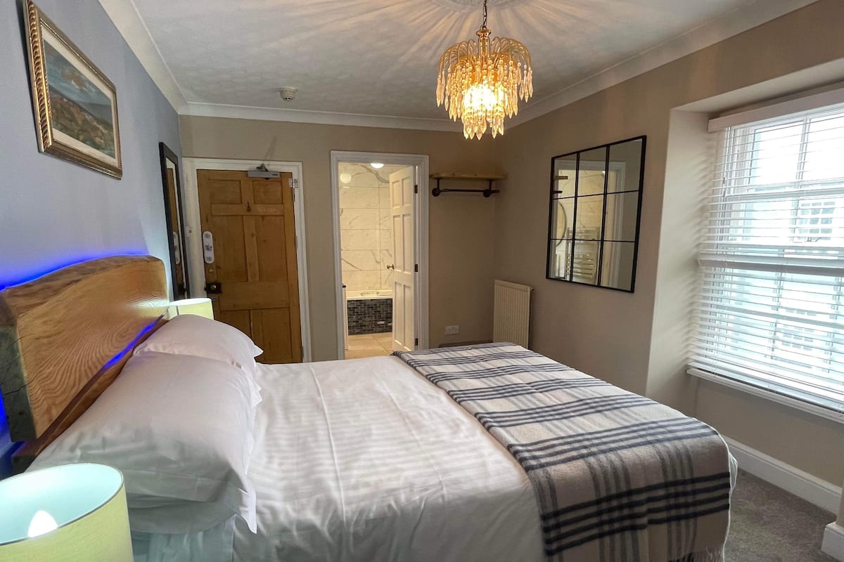 Une chambre à coucher avec un grand lit, une tête de lit en bois, un lustre, une fenêtre avec des stores et une porte donnant sur une salle de bain.