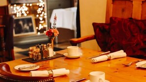 Mesa de jantar posta com pratos brancos, xícaras e talheres, um vaso com flores e saleiro e pimenteiro.