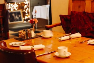 Una mesa de comedor dispuesta con platos blancos, tazas y cubiertos, un florero con flores, y saleros y pimenteros.
