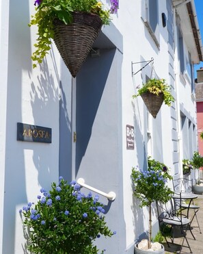 A building with a sign reading "AROSEA" and another sign indicating a bar. There are hanging baskets with plants and a small tree with blue flowers.