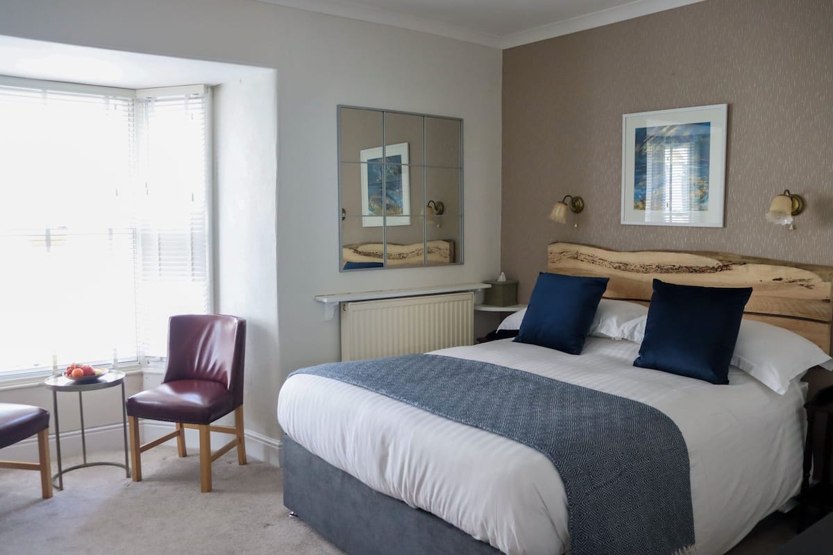 Une chambre à coucher avec un grand lit, une chaise, une petite table avec des fruits, un miroir et des œuvres d’art accrochées au mur.