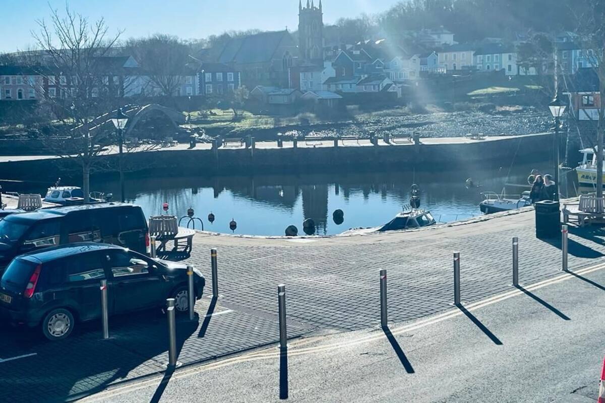 Un parking situé au bord de l’eau, avec une rivière paisible, une église en arrière-plan et un ciel dégagé.