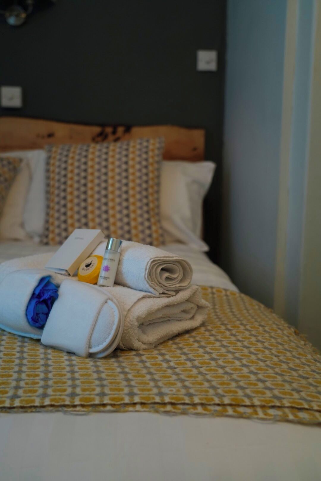 A neatly made bed with rolled towels, a bottle of lotion, and a folded garment on a patterned bedspread.