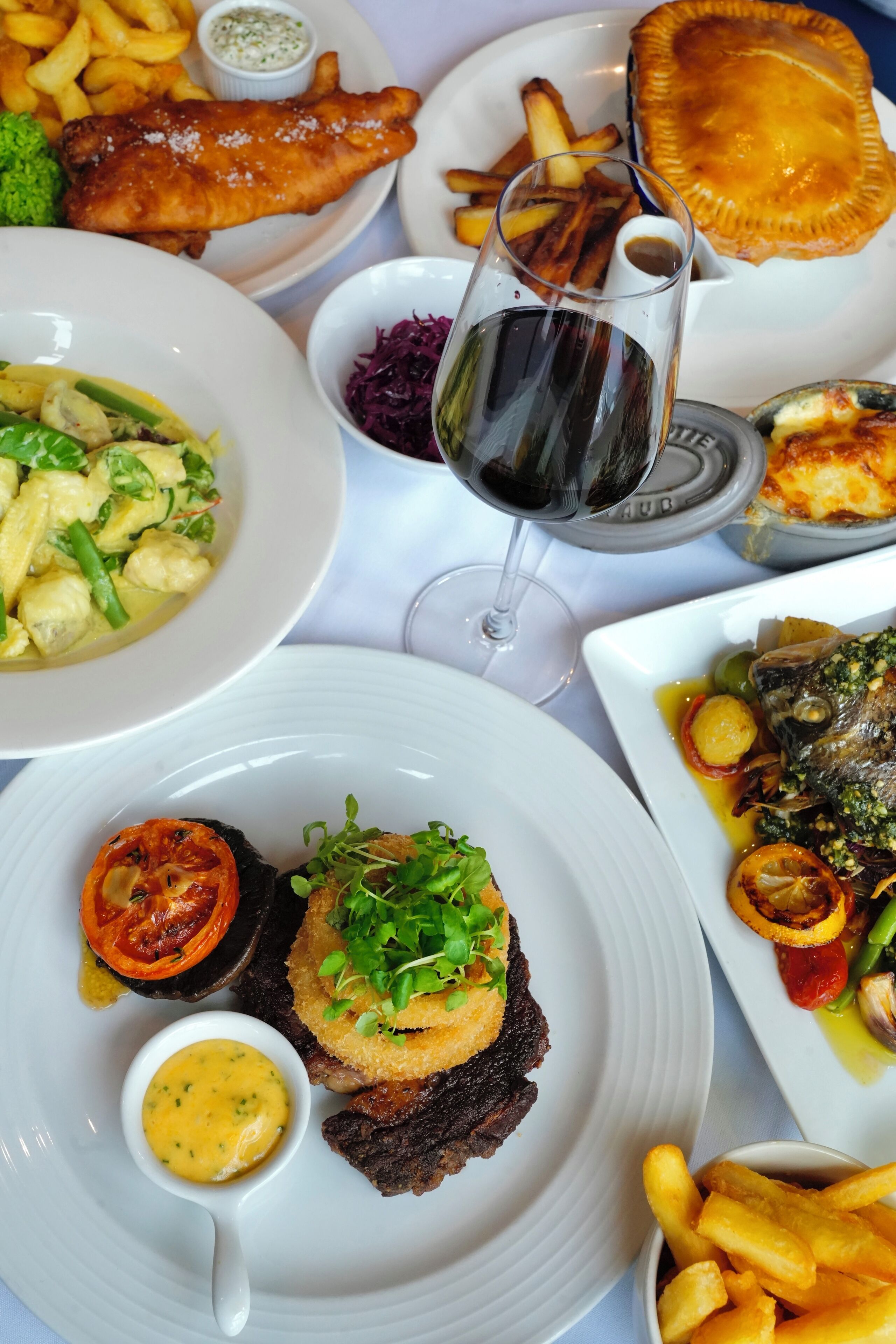 A variety of dishes including fish and chips, a pie, and a glass of red wine on a table.