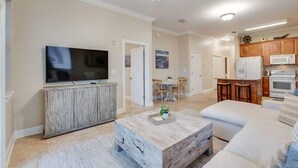A living room with a TV, wooden coffee table, and a sofa. There is a kitchen area with cabinets and appliances. - 3BR condo near beach w/ screened patio (Santa Rosa Beach)
