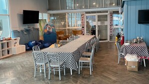 Ein geräumiger Essbereich mit einem langen Tisch, gedeckt für mehrere Personen, umgeben von Stühlen. Es gibt Deckenventilatoren, einen an der Wand montierten Fernseher und ein Schild mit der Aufschrift 'WELCOME'.