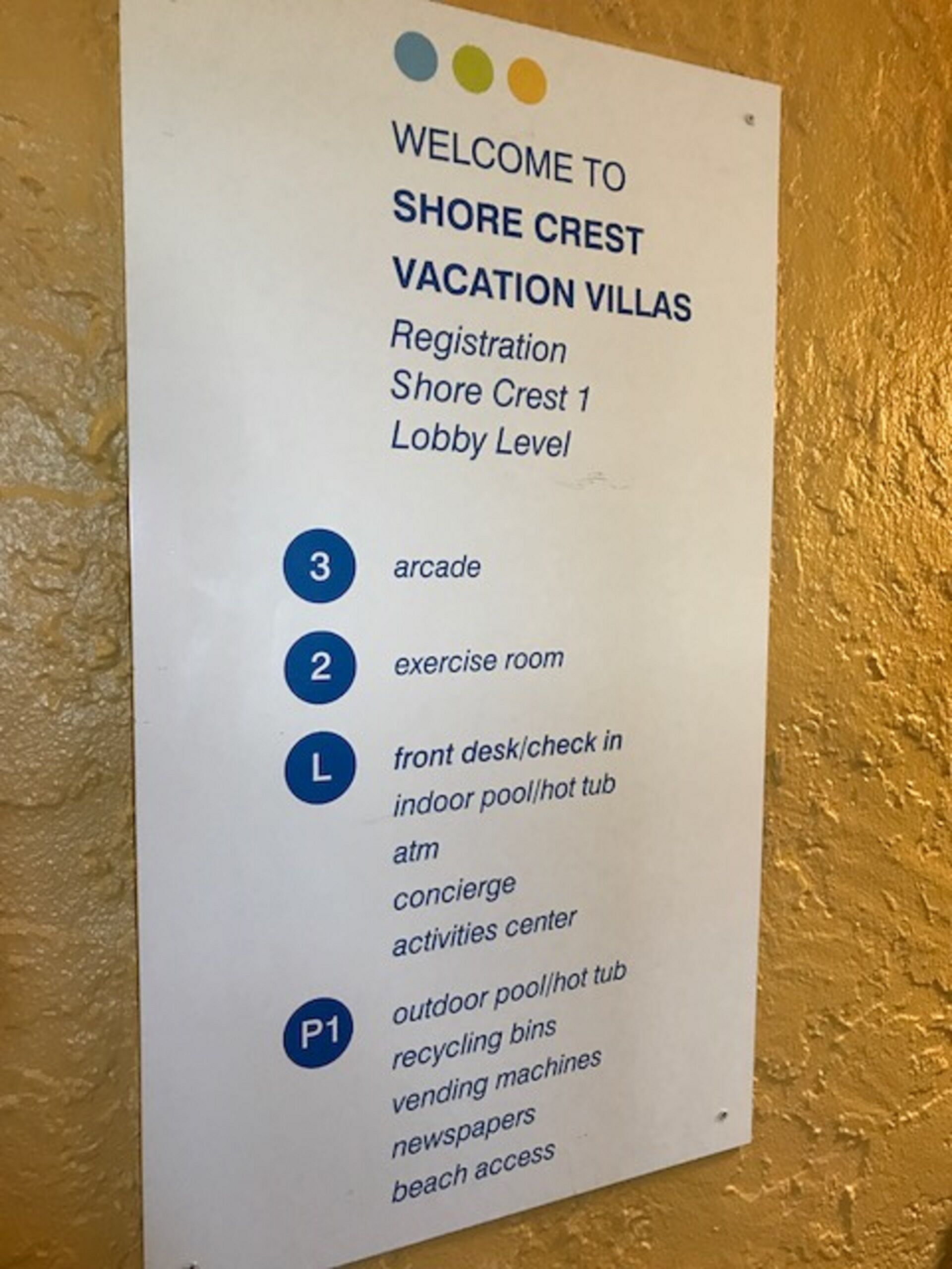 Schild für Shore Crest Vacation Villas mit Auflistung der Annehmlichkeiten wie Rezeption, Lobbyebene, Spielhalle, Fitnessraum, Rezeption, Hallenbad, Außenpool, Concierge-Service, Aktivitätszentrum, Recyclingbehälter, Getränkeautomaten, Zeitschriften und Zugang zum Strand.