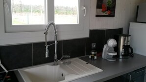 A modern kitchen with a white sink, a stainless steel faucet, a coffee maker, and a window with a view of trees. - Chalet 4 people with shared pool (Hourtin)