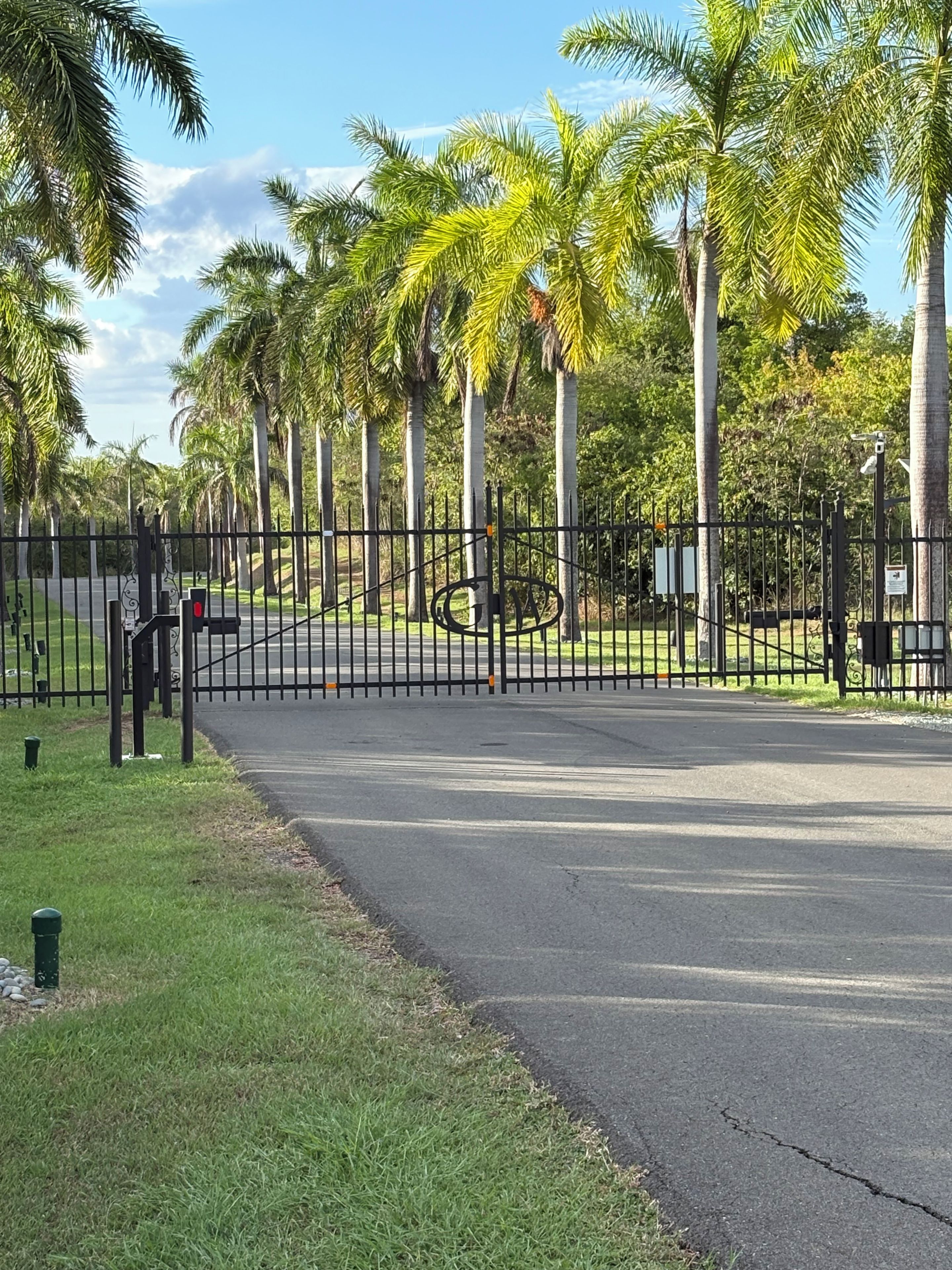 Una entrada con portón y cerca de metal, un sendero peatonal y palmeras al fondo.