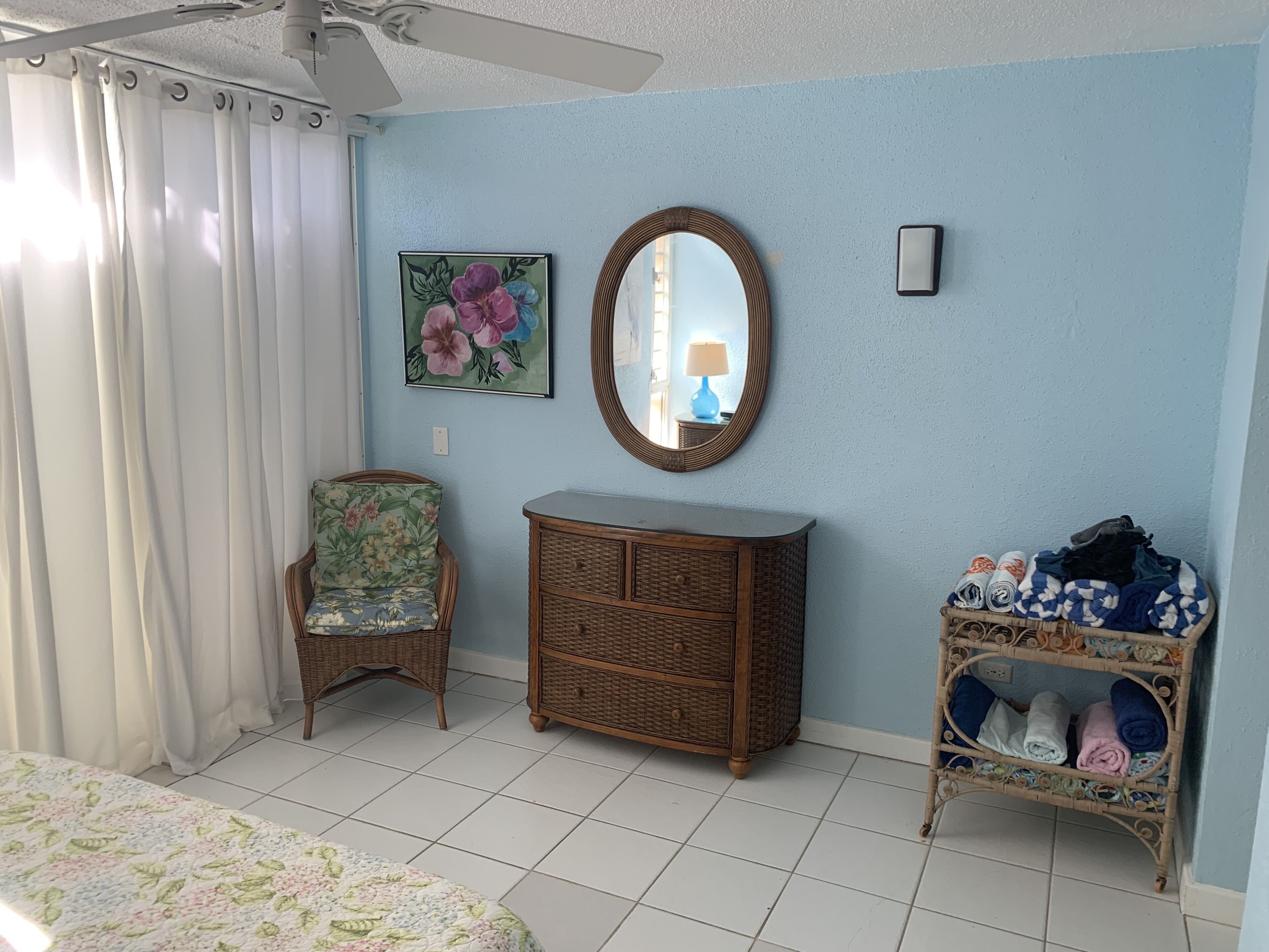 Un dormitorio con una cama, un aparador de mimbre, una silla, un espejo y una estantería con ropa doblada.