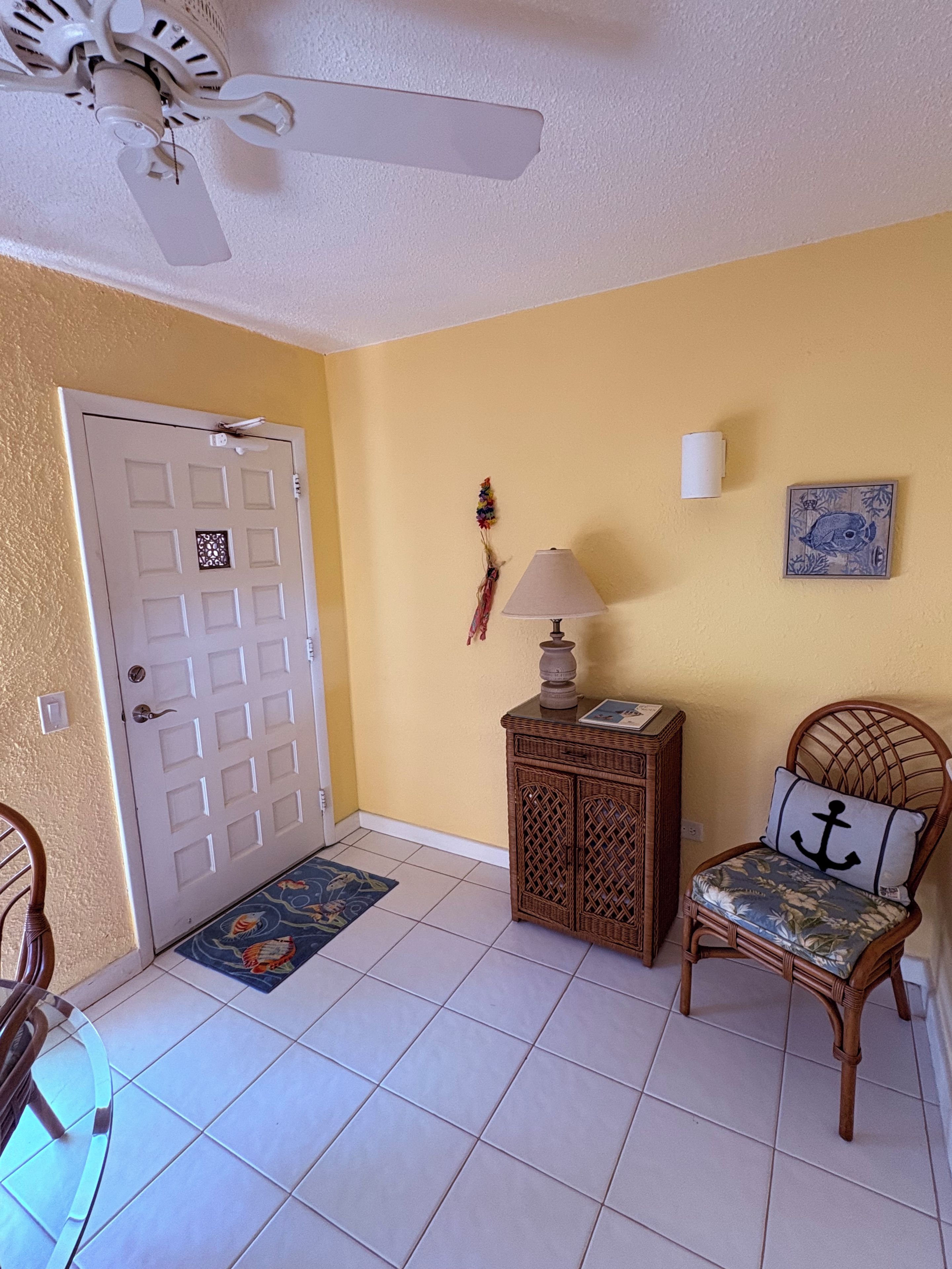Una habitación con una puerta blanca, un ventilador de techo, una pequeña mesa con lámpara, una silla de mimbre con cojín y un adorno en la pared.
