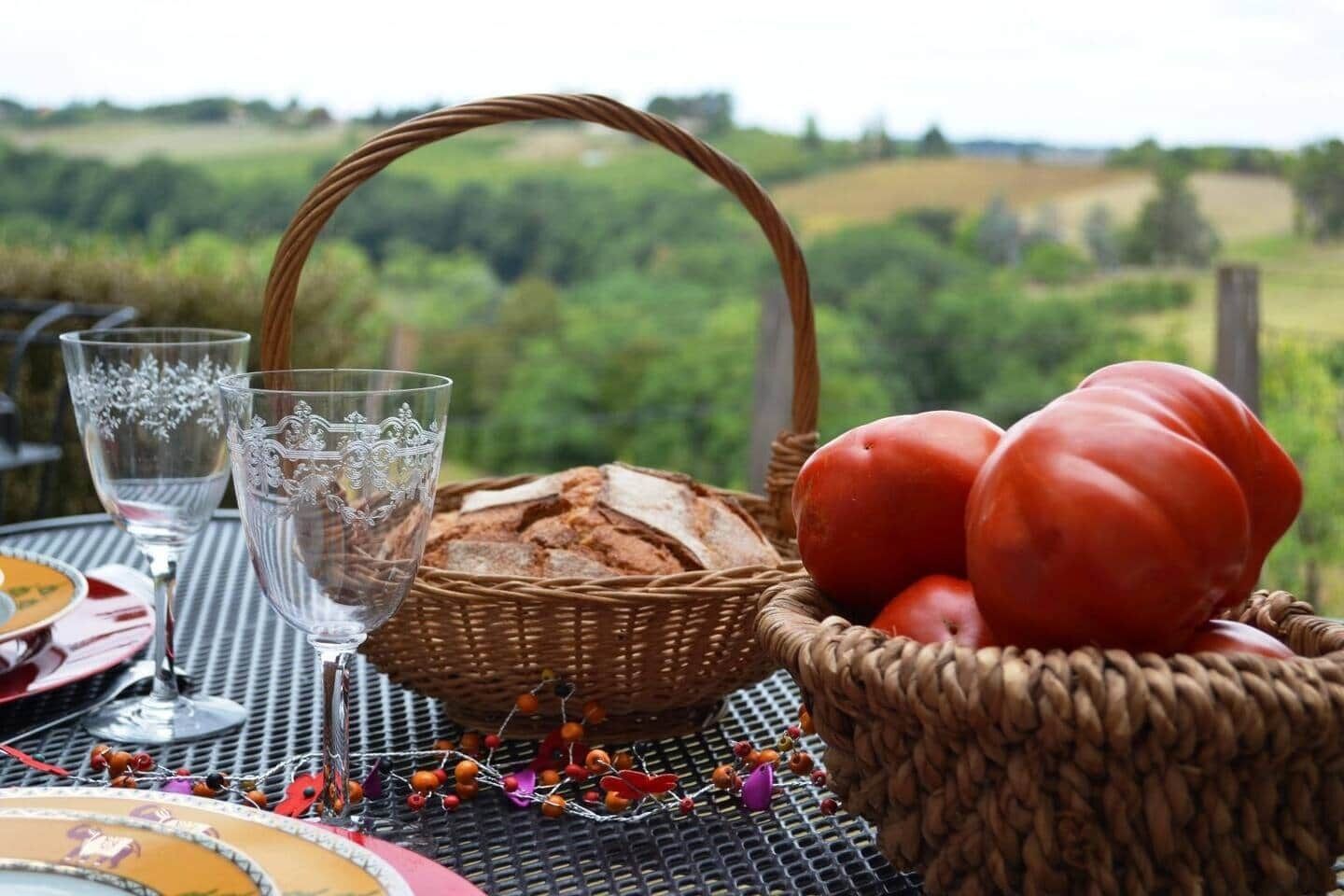 A table set with a basket of bread, tomatoes, and decorative elements, with a scenic view in the background.