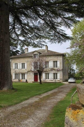 A two-story stone building with a red door, a gravel driveway, and a tree in the foreground. - To complete (Madaillan)