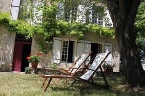A rustic building with a stone facade, a red door, and a large tree in front. There are two wooden chairs and a potted plant with red flowers. - To complete (Madaillan)