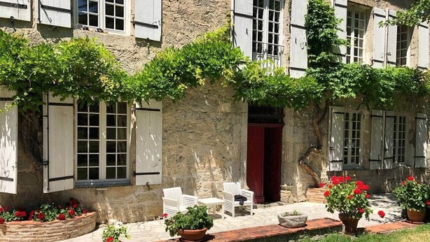 A rustic stone building with white shutters, a red door, and outdoor seating.