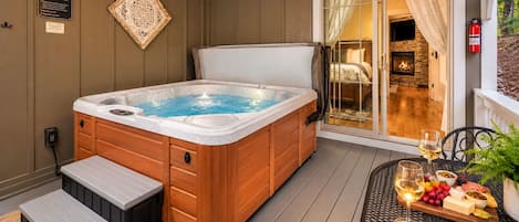 A hot tub with wooden steps and a wooden cabinet, a patio table with a glass of wine and fruit, and a stone fireplace.