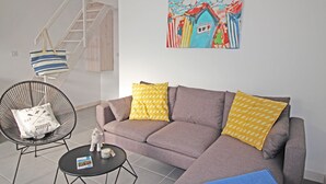 A living room with a grey sofa, two yellow patterned pillows, a black metal chair, a small white cushion with text, a round black table with a book and a candle, and a colorful painting on the wall. - Le Clos des Flots, The Fleet - House "L'Océan" - 2 bedrooms, private patio (La Flotte)
