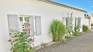 A white building with grey shutters, a gravel driveway, and various plants including pink flowers and tall green grass. - Le Clos des Flots, The Fleet - House "L'Océan" - 2 bedrooms, private patio (La Flotte)