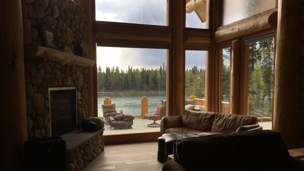 A cozy cabin interior with a stone fireplace, a leather sofa, and a view of a lake and forest through large windows.