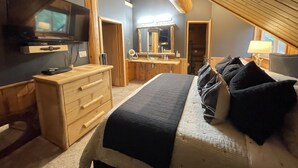 A bedroom with a bed, dresser, vanity, and a television.