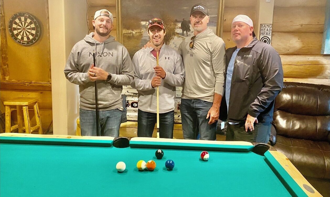 A group of people standing around a pool table in a room with wooden walls and a dartboard.