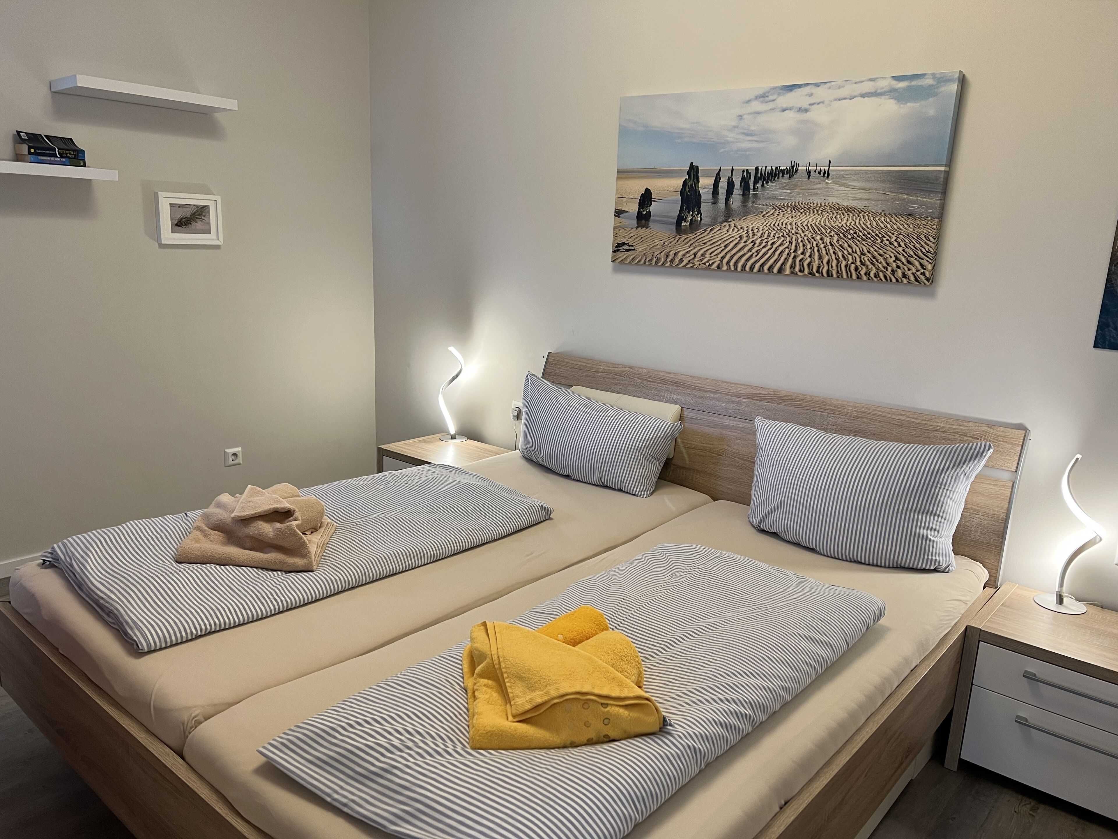 A double bed with striped bedding, two bedside tables with lamps, a painting of a beach scene, and a small shelf with books and a framed picture.