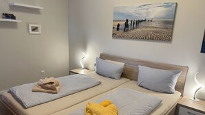 A double bed with striped bedding, two bedside tables with lamps, a painting of a beach scene, and a small shelf with books and a framed picture.