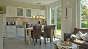 A modern kitchen with white cabinets, a dining area with wooden chairs, and a sofa with a view of greenery outside.