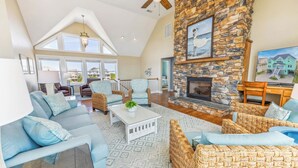 A spacious living room with a stone fireplace, comfortable seating, and large windows offering views of the surrounding area. - Premier Oceanfront Dog Friendly Home w/ Pool, Hot Tub, EV Charger, and Game Room (Hatteras)