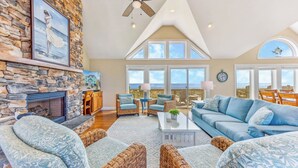 A spacious living room with a stone fireplace, wicker furniture, and large windows offering ocean views. - Premier Oceanfront Dog Friendly Home w/ Pool, Hot Tub, EV Charger, and Game Room (Hatteras)