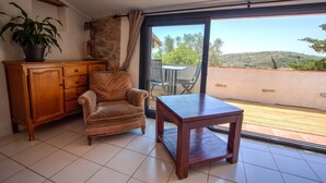 A room with a wooden cabinet, a cushioned chair, a small table, and a sliding glass door leading to an outdoor area with a view of hills.