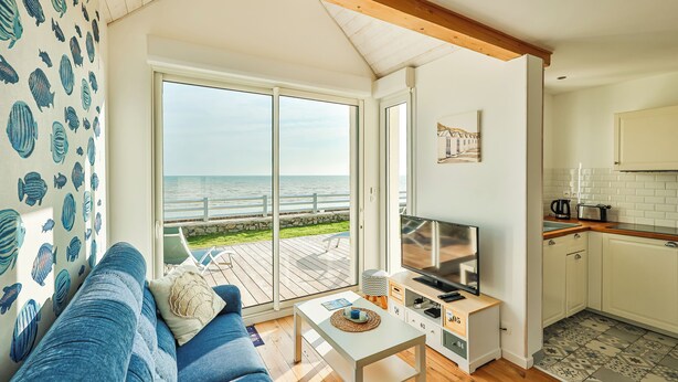 A living room with a large sliding glass door leading to a balcony with a view of the sea. There is a TV on a white stand, a blue sofa, and a small table with a remote control.